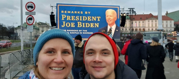 Kolega i koleżanka pozują do zdjęcia lekko uśmiechnięci w zimowych ubraniach. W oddali duży baner z niebieskim tłem z Joe Bidenem.