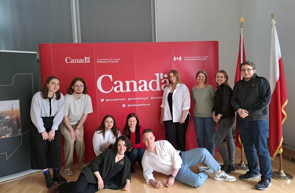 Grupa studentów i studentek pozujący do zdjęcia. Dwójka kuca, dwójka leży. W tle czerwony ekran z napisem „Canada”.