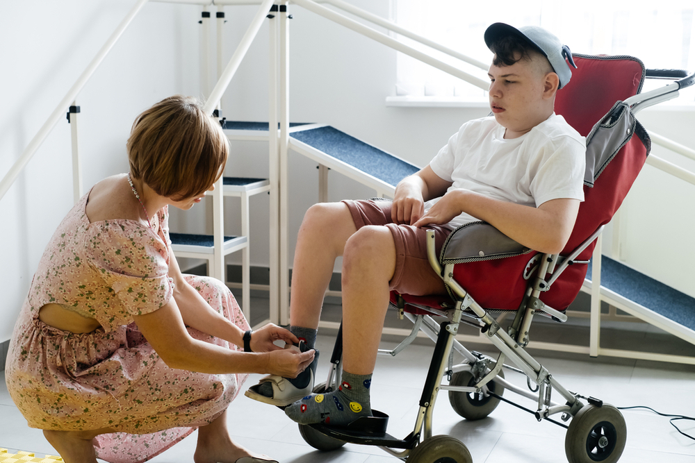 A mother helps a 17-year-old disabled boy with cerebral palsy in a wheelchair put on his shoes before going for a walk.